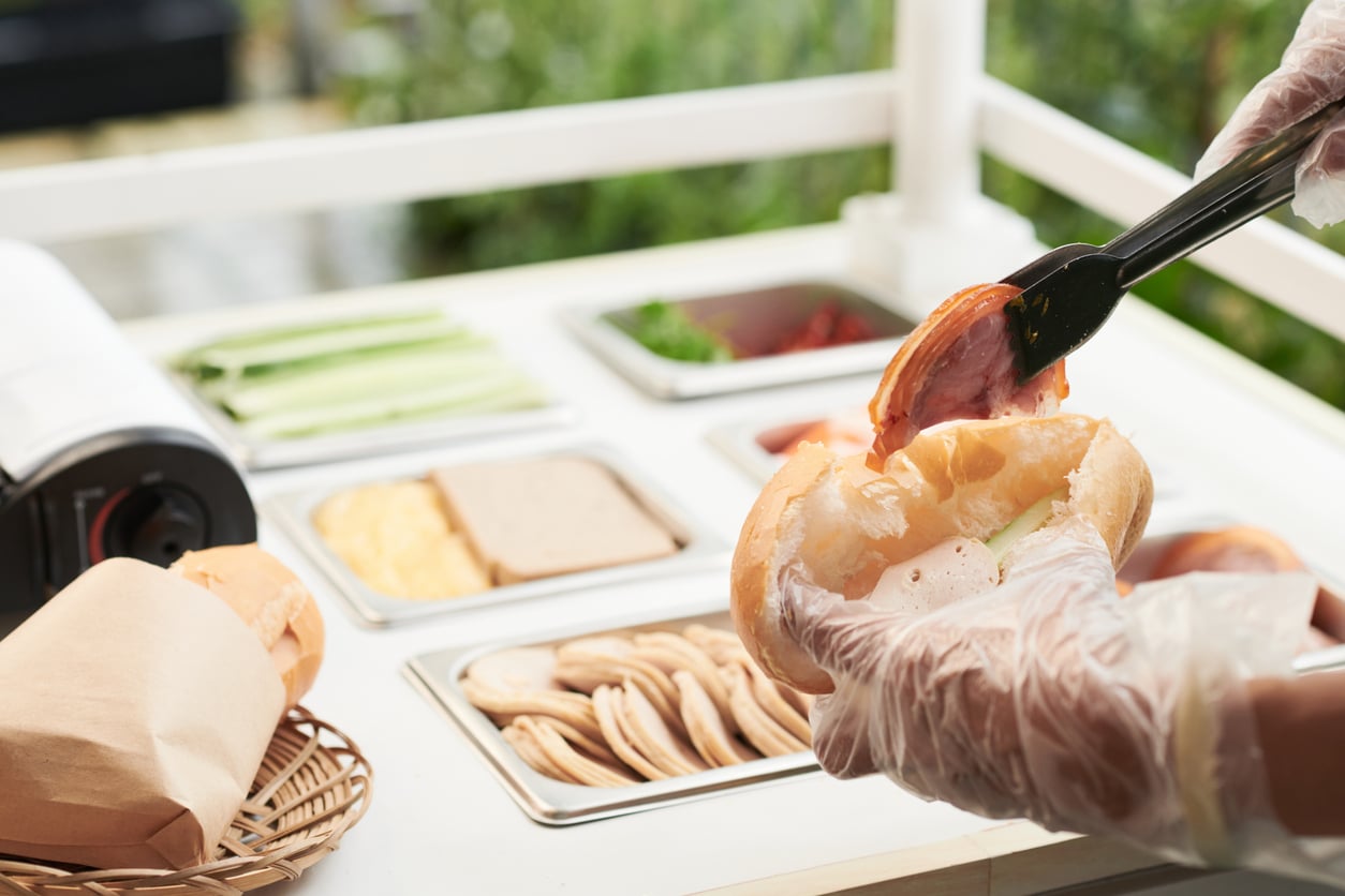 A person wearing plastic gloves uses tongs to place a slice of meat into a sandwich bun at a food preparation station.