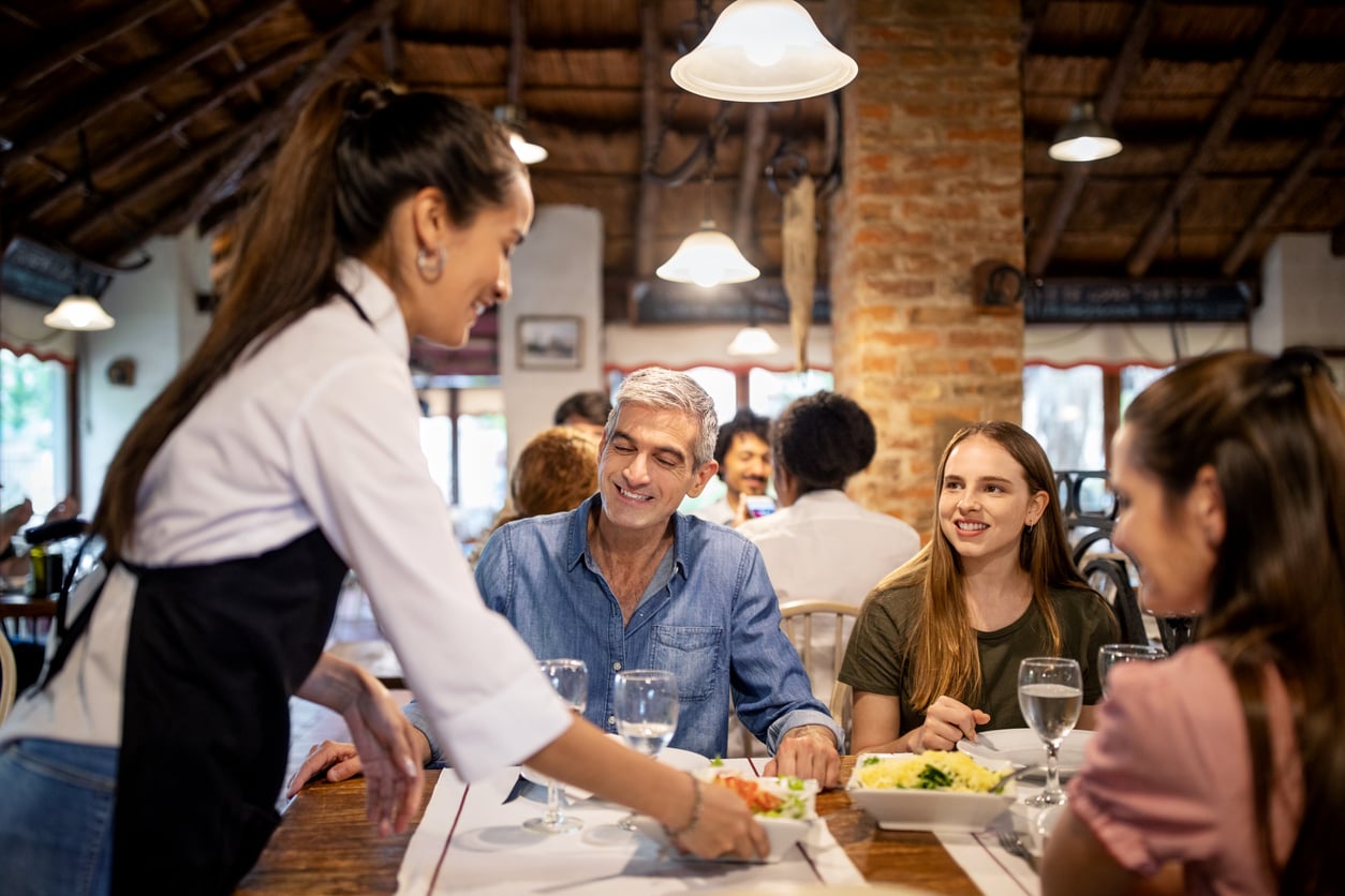 A smiling server places a plate of food from the restaurant menu on a table in front of a group of people dining.