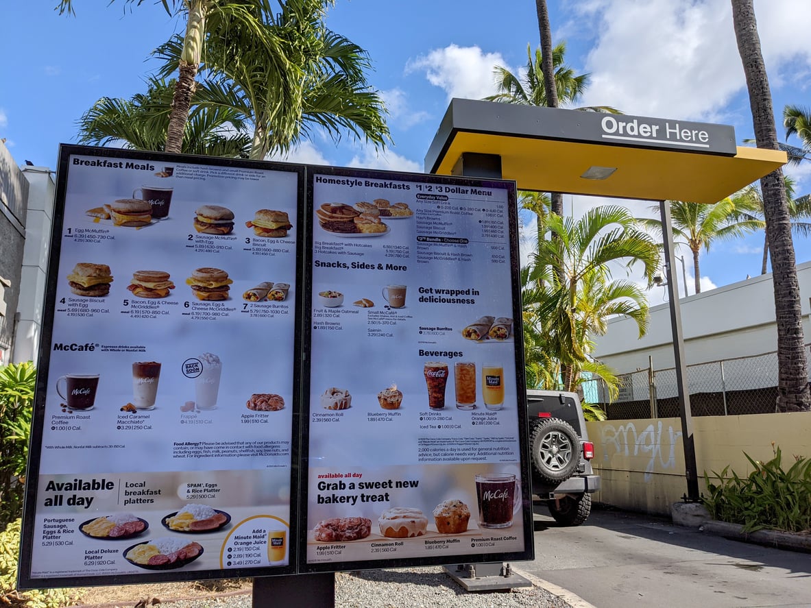 A McDonald’s drive-thru menu board displaying breakfast, snack, and beverage options with prices and calorie counts.