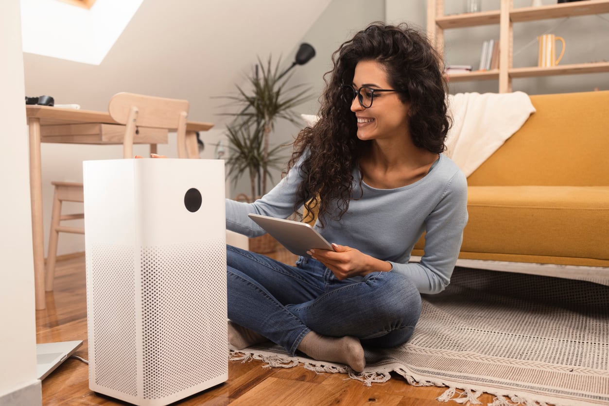 A smiling woman sitting on the floor uses a tablet to control an air purifier.