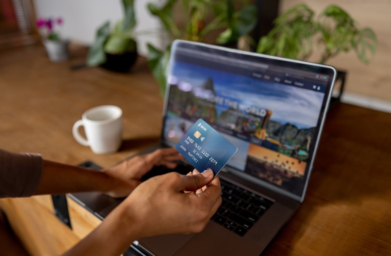 A person holding a blue credit card while sitting in front of an open laptop on a wooden desk to make trip planning decisions.