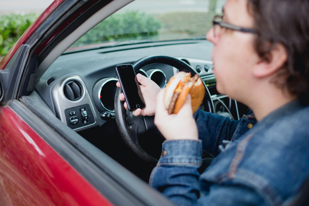 A person driving a car while holding a smartphone and a burger.