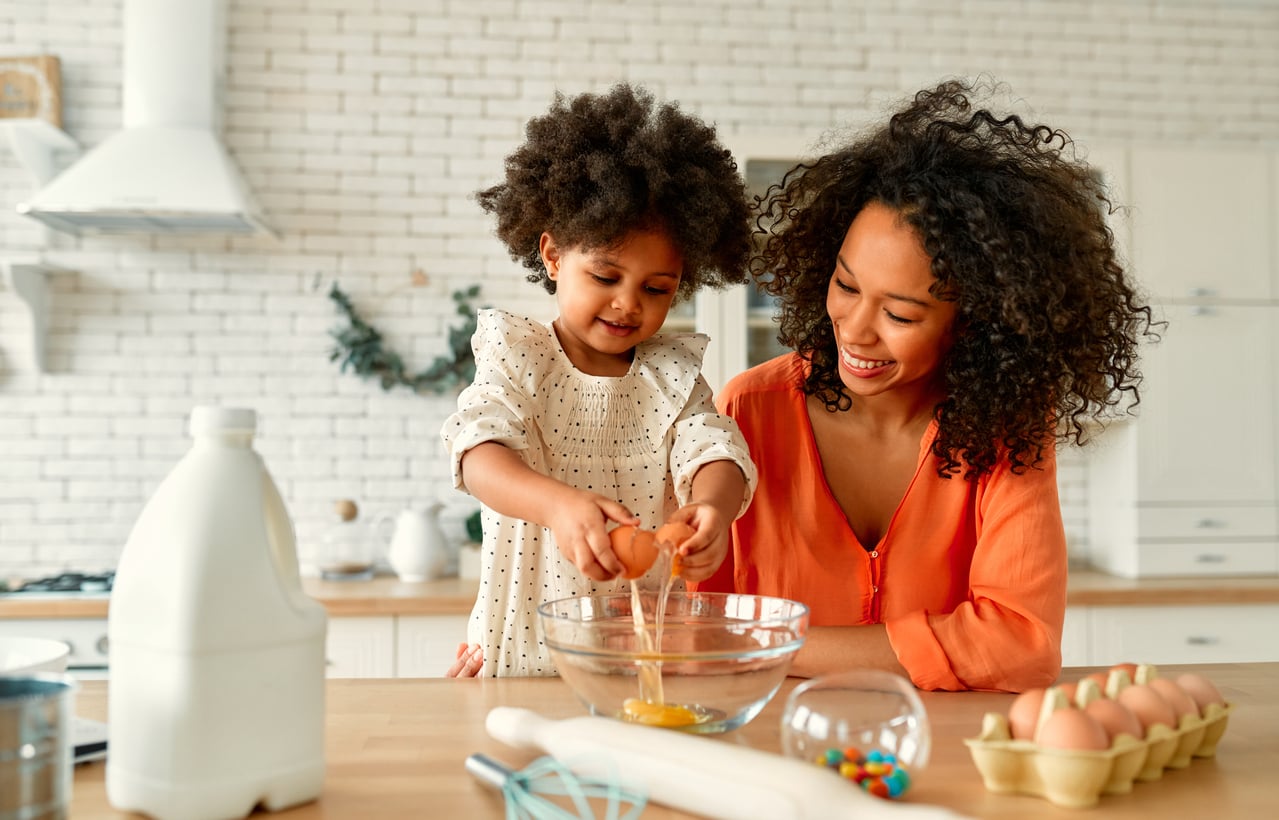 A woman watches a young child crack an egg into a glass bowl on a kitchen counter.