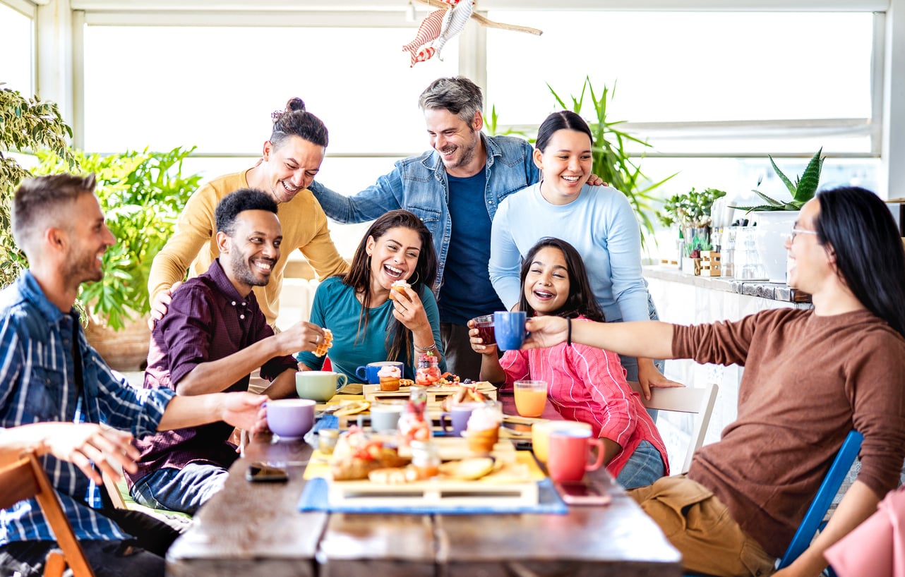 A diverse group of seven friends laughing and talking while sitting around a wooden table set with drinks and snacks.