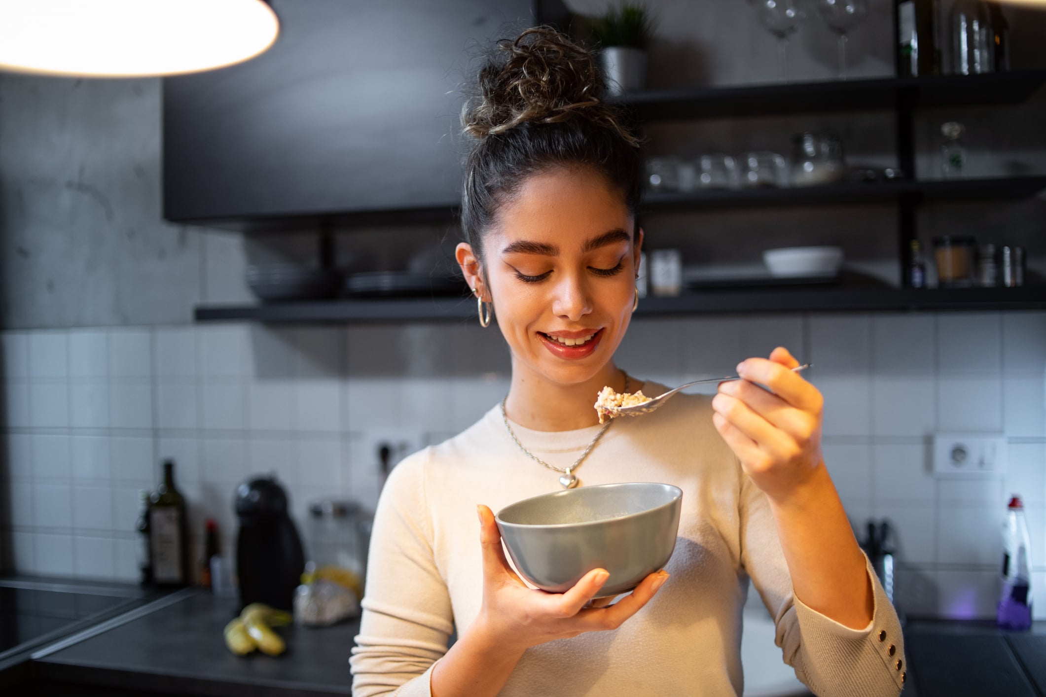 A smiling woman in a kitchen holds a bowl of breakfast foods and lifts a spoonful to her mouth.