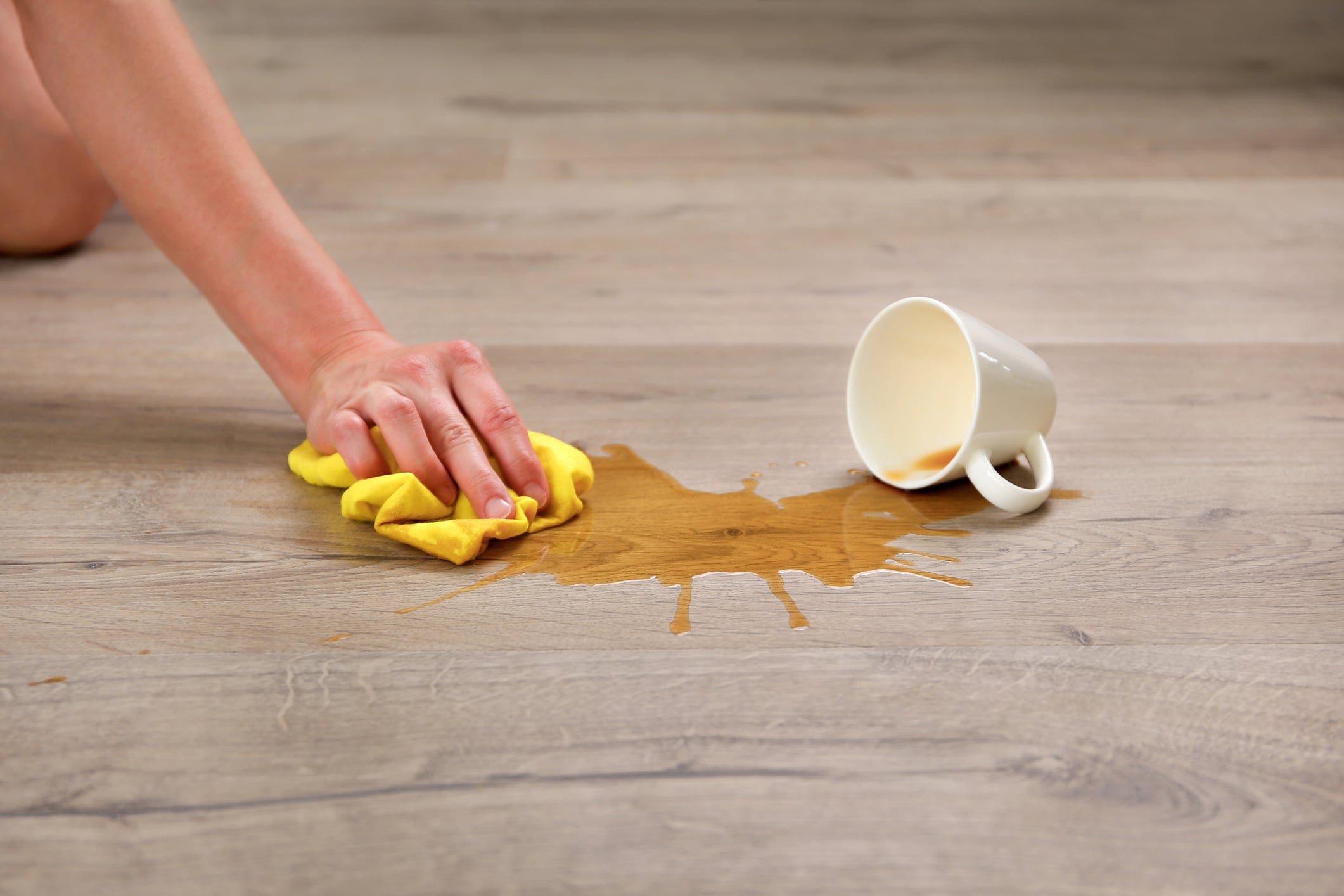 A person wipes up a coffee spill from a light wood floor with a yellow cloth near an overturned white mug, a quick task for home improvements.