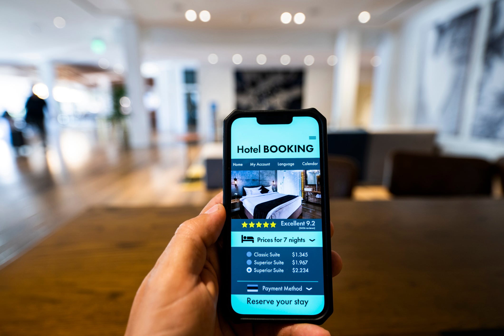 A hand holds a smartphone displaying a hotel booking app with room options and prices for a seven-night stay.