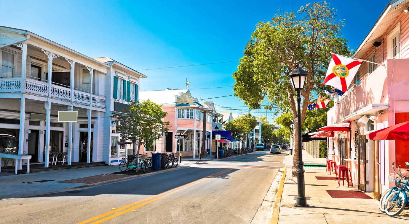 A sunny street in Key West lined with pastel-colored buildings, bicycles, and the Florida state flag hanging from a porch.
