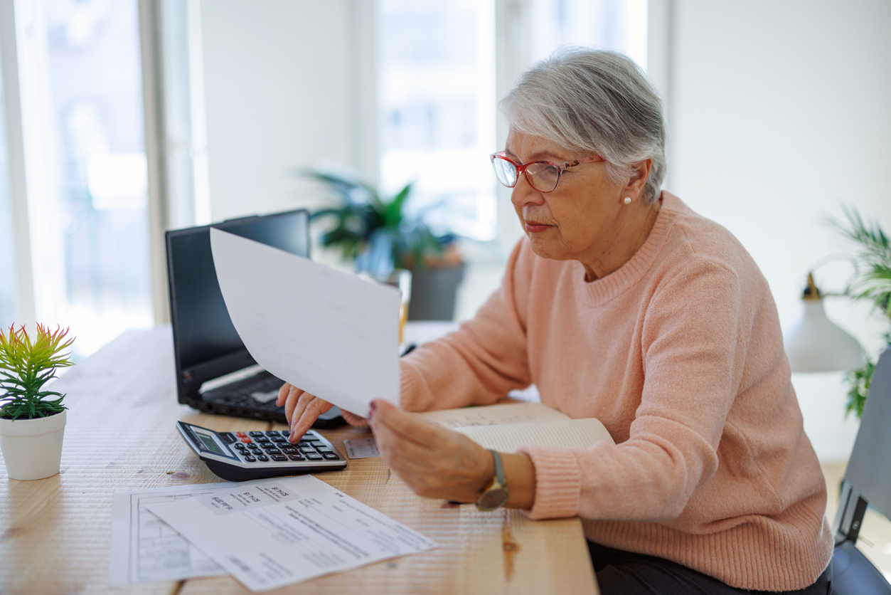 An older woman in a pink sweater sits at a table, using a calculator and looking at a document.