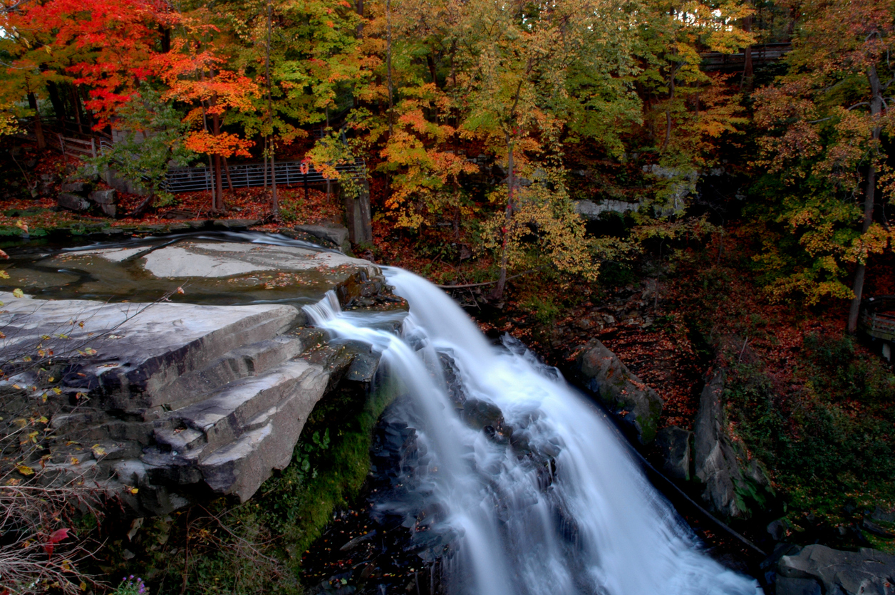A waterfall cascades over a rocky ledge surrounded by trees with vibrant autumn foliage, a scenic view for places to hike in the fall.