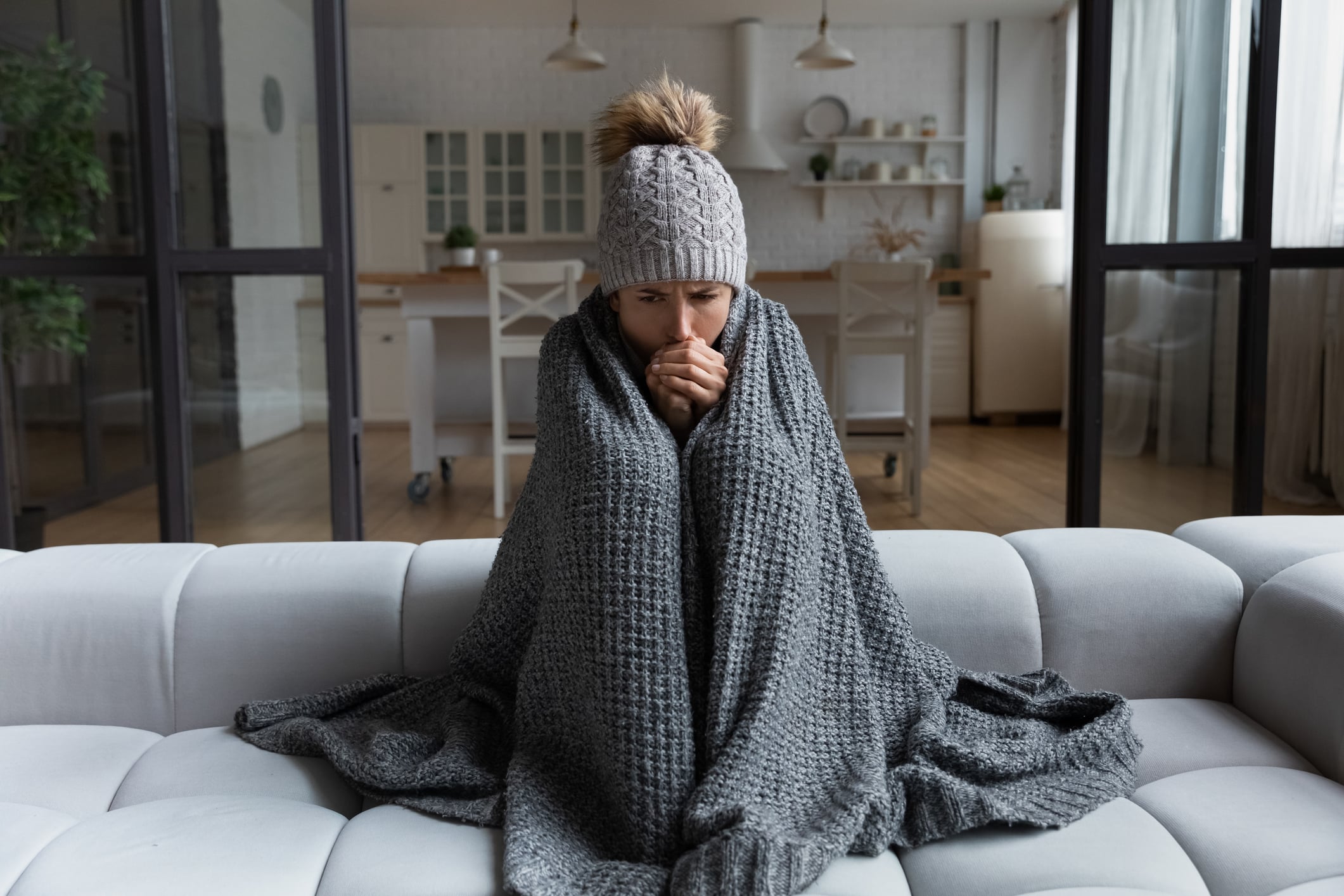 A woman sits on a sofa in a home requiring improvements, wrapped in a thick grey blanket and wearing a knit beanie, shivering with her hands held to her chin.