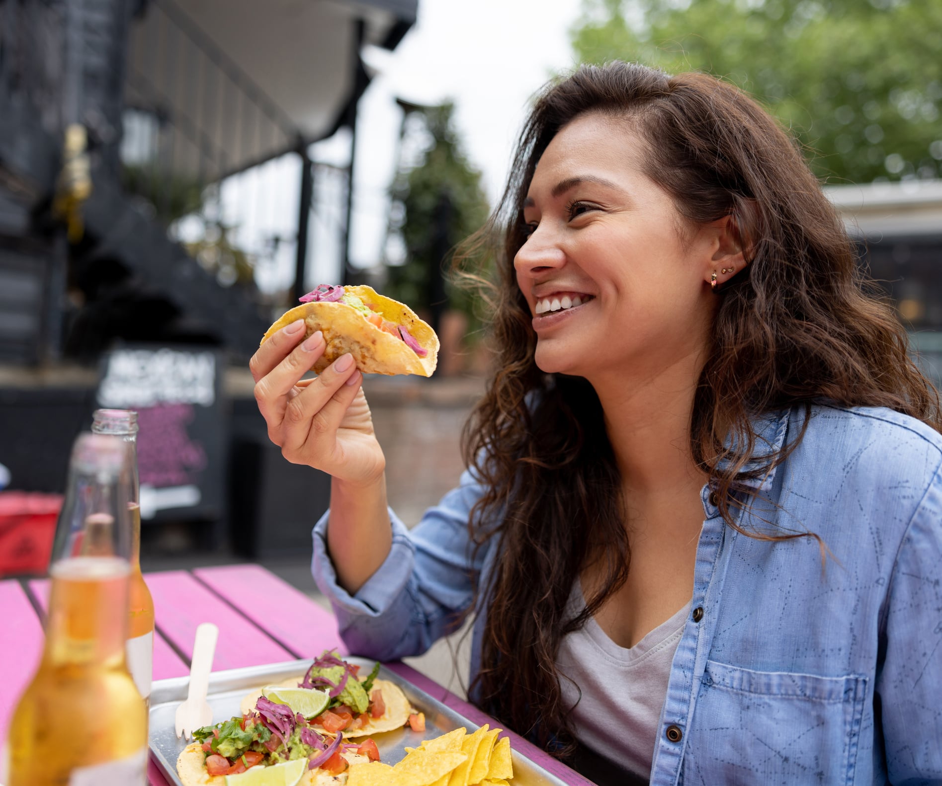 A smiling woman eating breakfast tacos at an outdoor table with a tray of food and drinks.