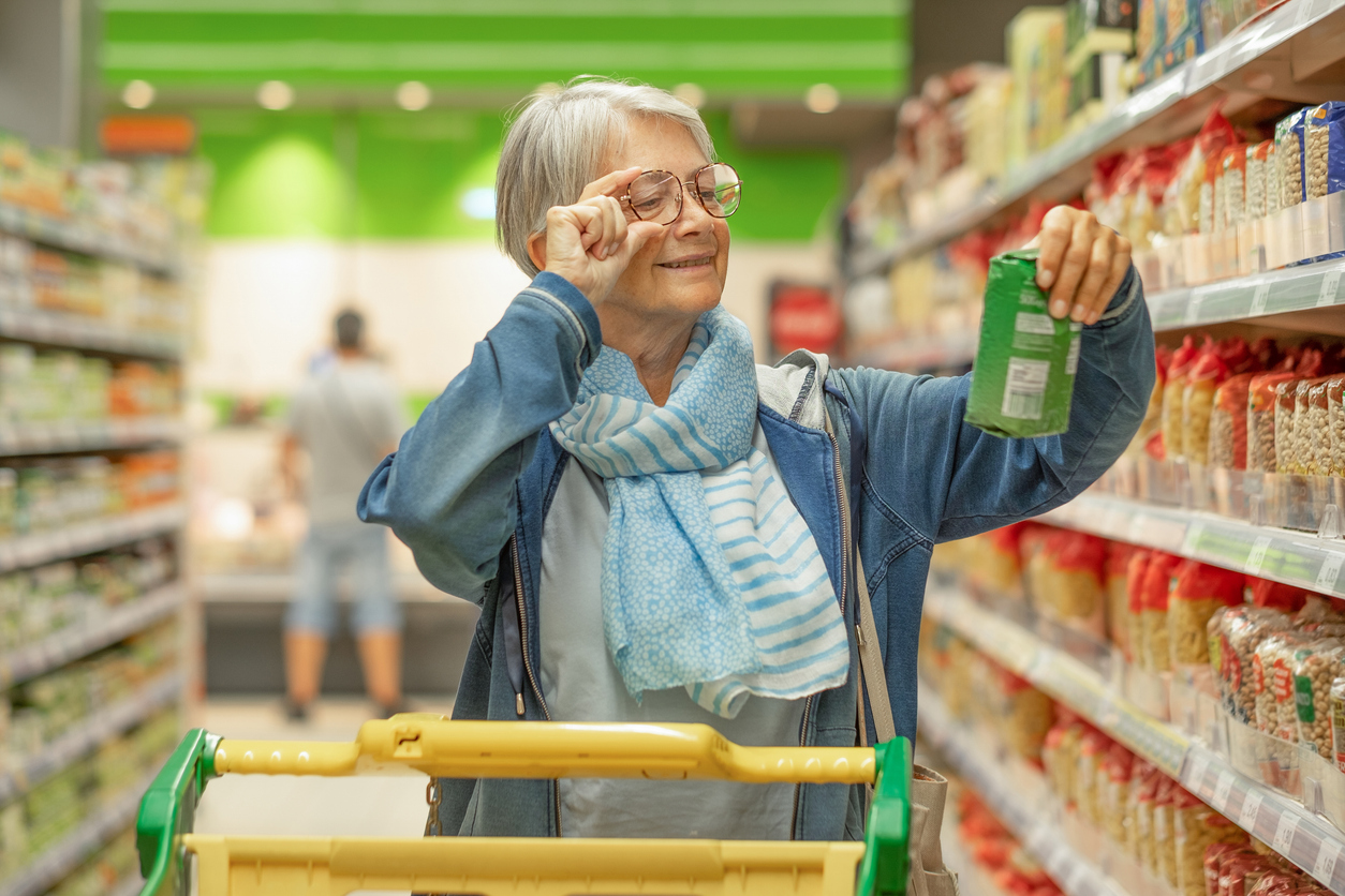 An older woman with glasses adjusting them while examining a green product package in a grocery store aisle.