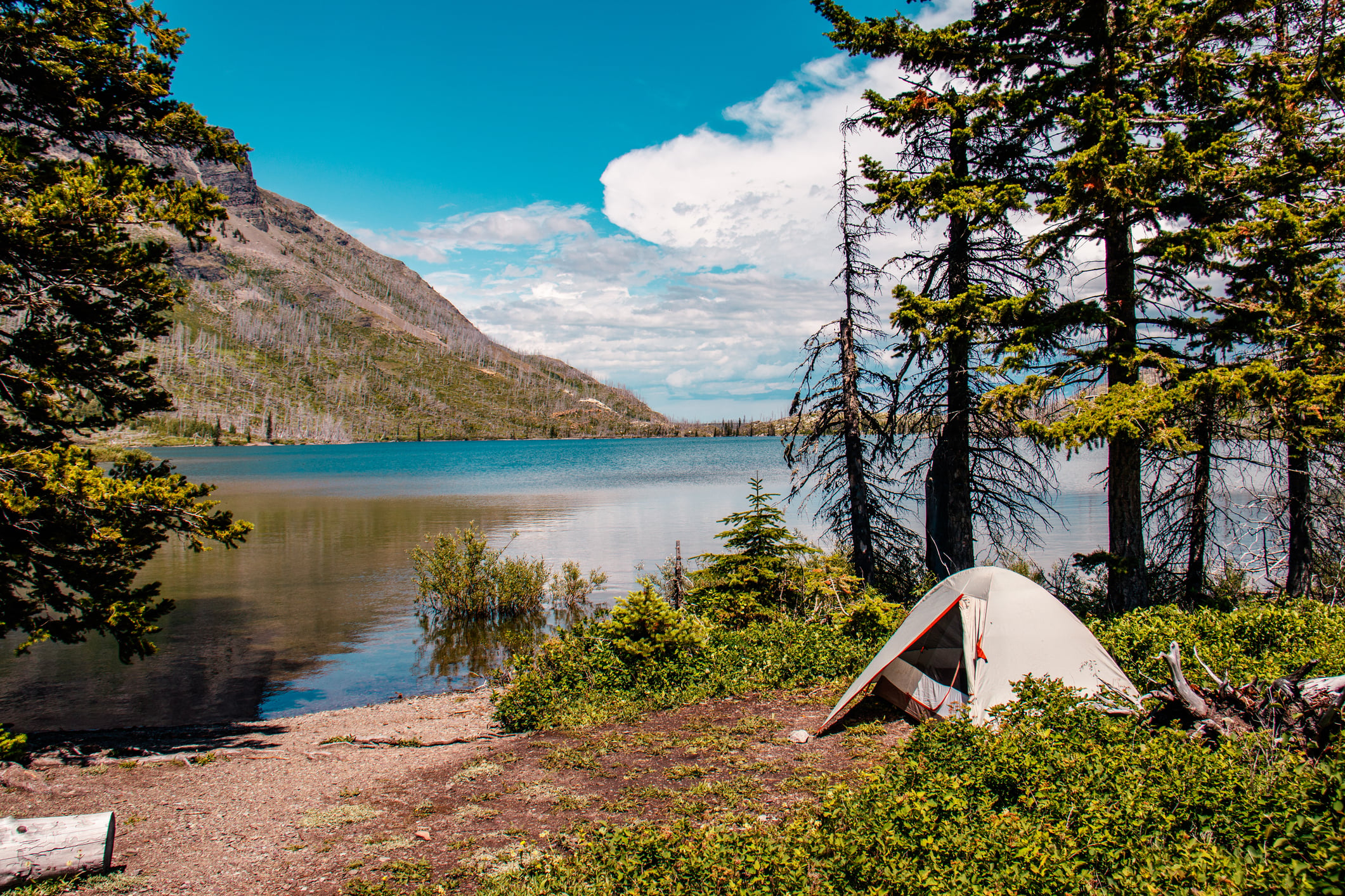 A white camping tent set up on the shore of a lake with mountains in the background.