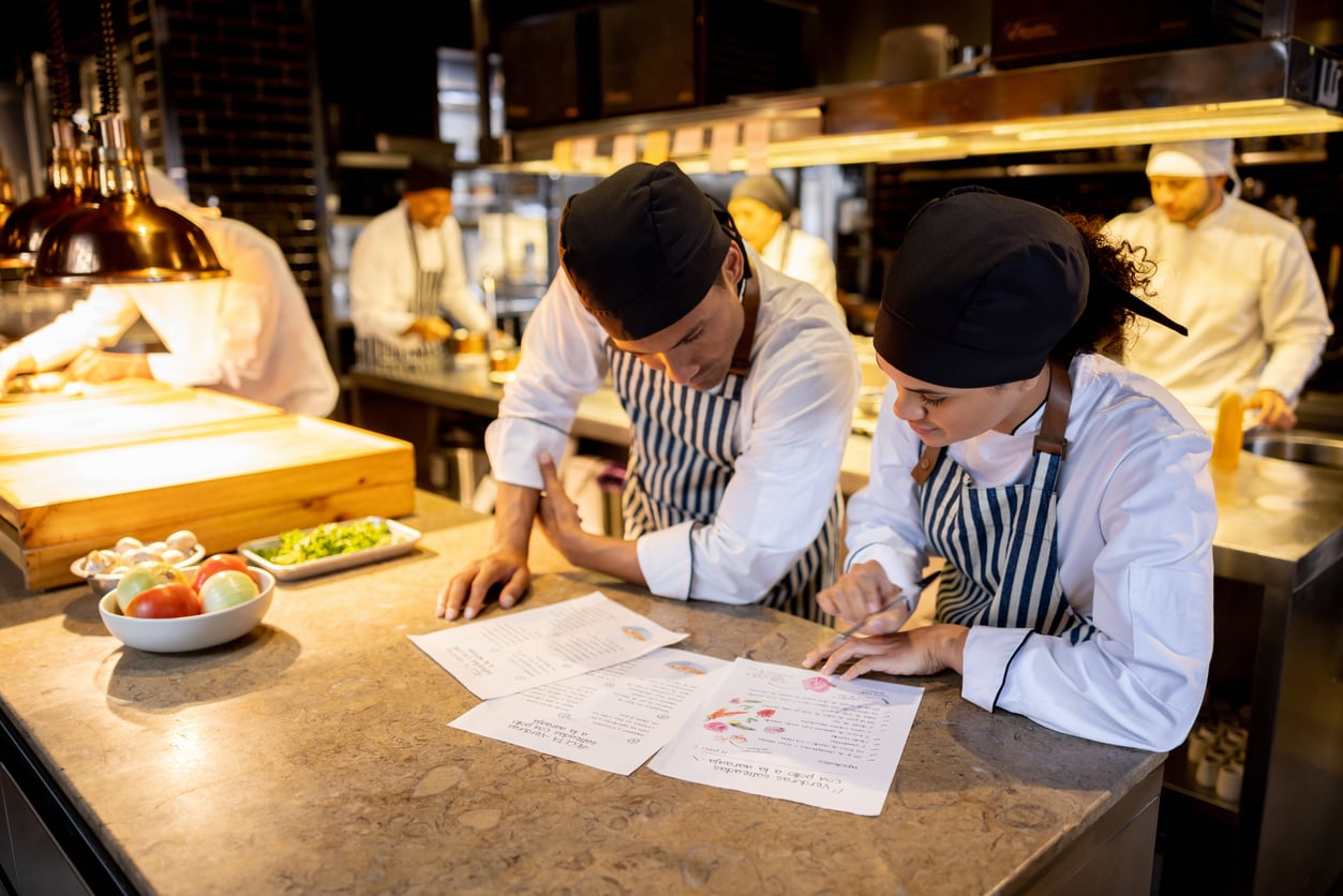 Two chefs in white uniforms and striped aprons review a printed restaurant menu on a kitchen counter, with other staff working in the background.