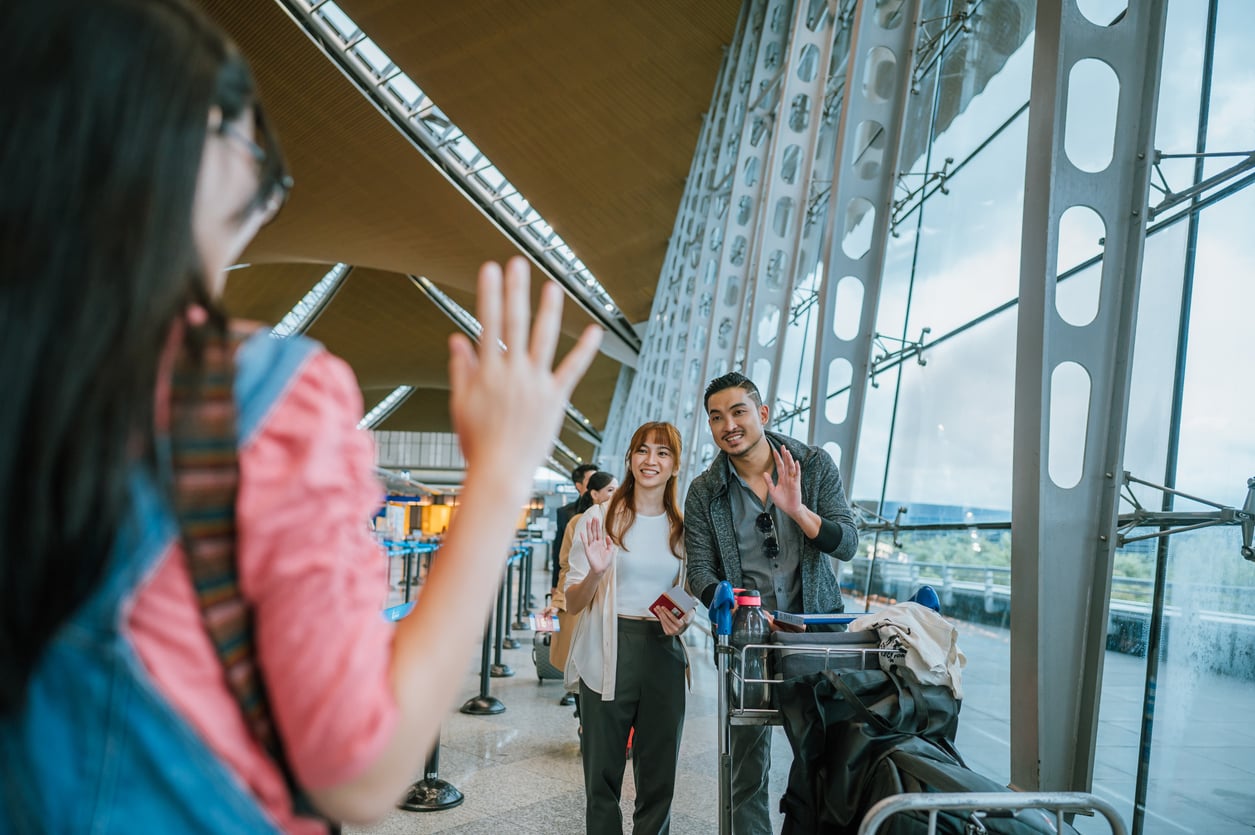 A person waves hello to a couple with luggage standing in an airport terminal.