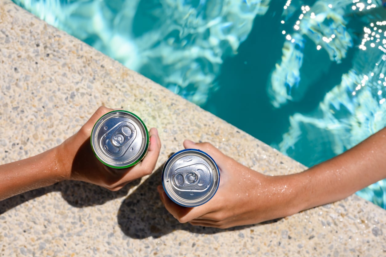 Two people holding beverage cans while resting their arms on the edge of a swimming pool.