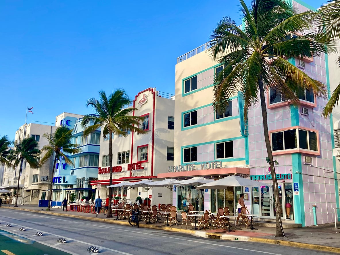A row of colorful art deco hotels along Ocean Drive in Miami Beach, featuring palm trees and outdoor sidewalk cafe seating.
