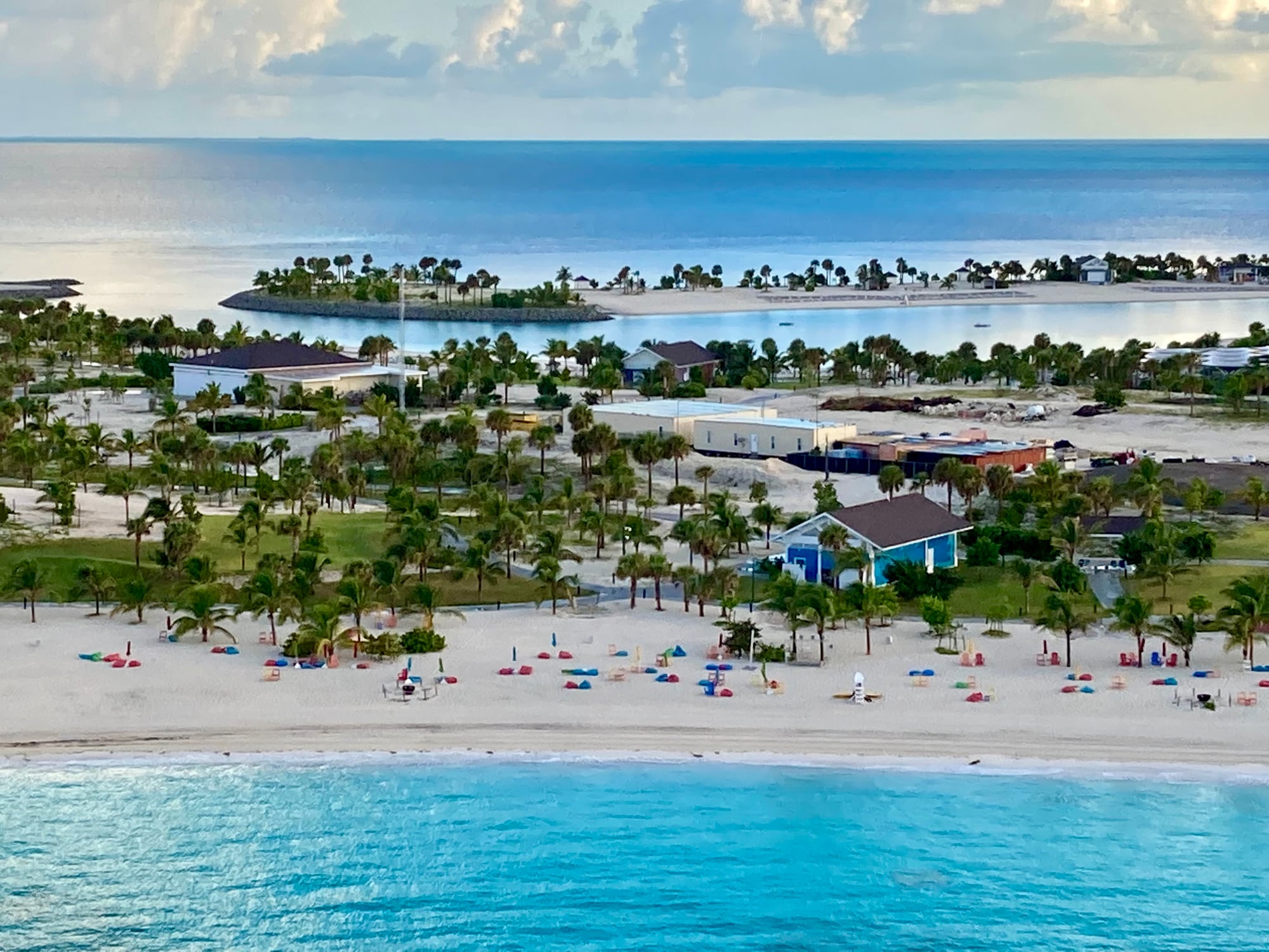 Aerial view of a tropical resort beach with rows of colorful lounge chairs, scattered palm trees, and coastal buildings overlooking a turquoise sea.