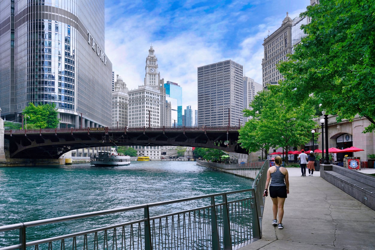 A person walks along the Chicago Riverwalk with the Chicago River, the Wabash Avenue bridge, and skyscrapers in the background.