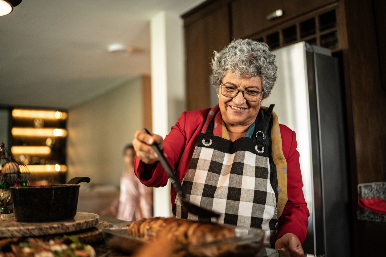 A smiling older woman with grey hair and an apron using a spatula to serve food from a glass baking dish in a kitchen.