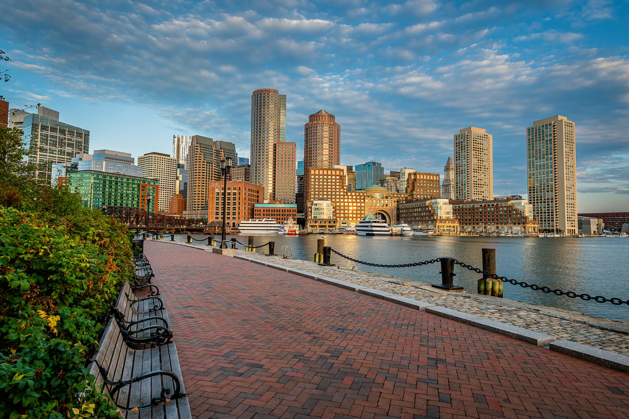 A brick walkway with empty benches along the water, looking across the harbor toward the Boston skyline at sunset.
