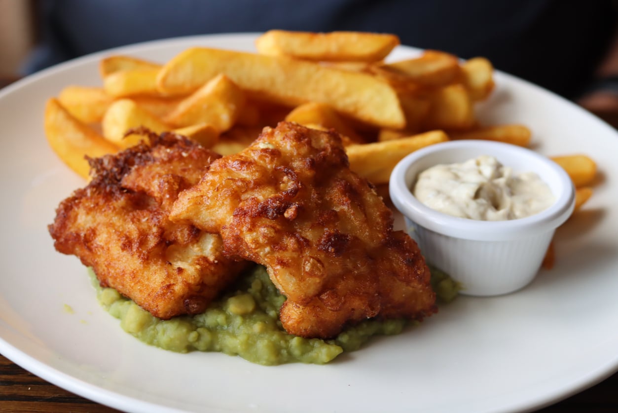 Two pieces of fried fish served over mushy peas with a side of thick-cut fries and a small dish of tartar sauce.