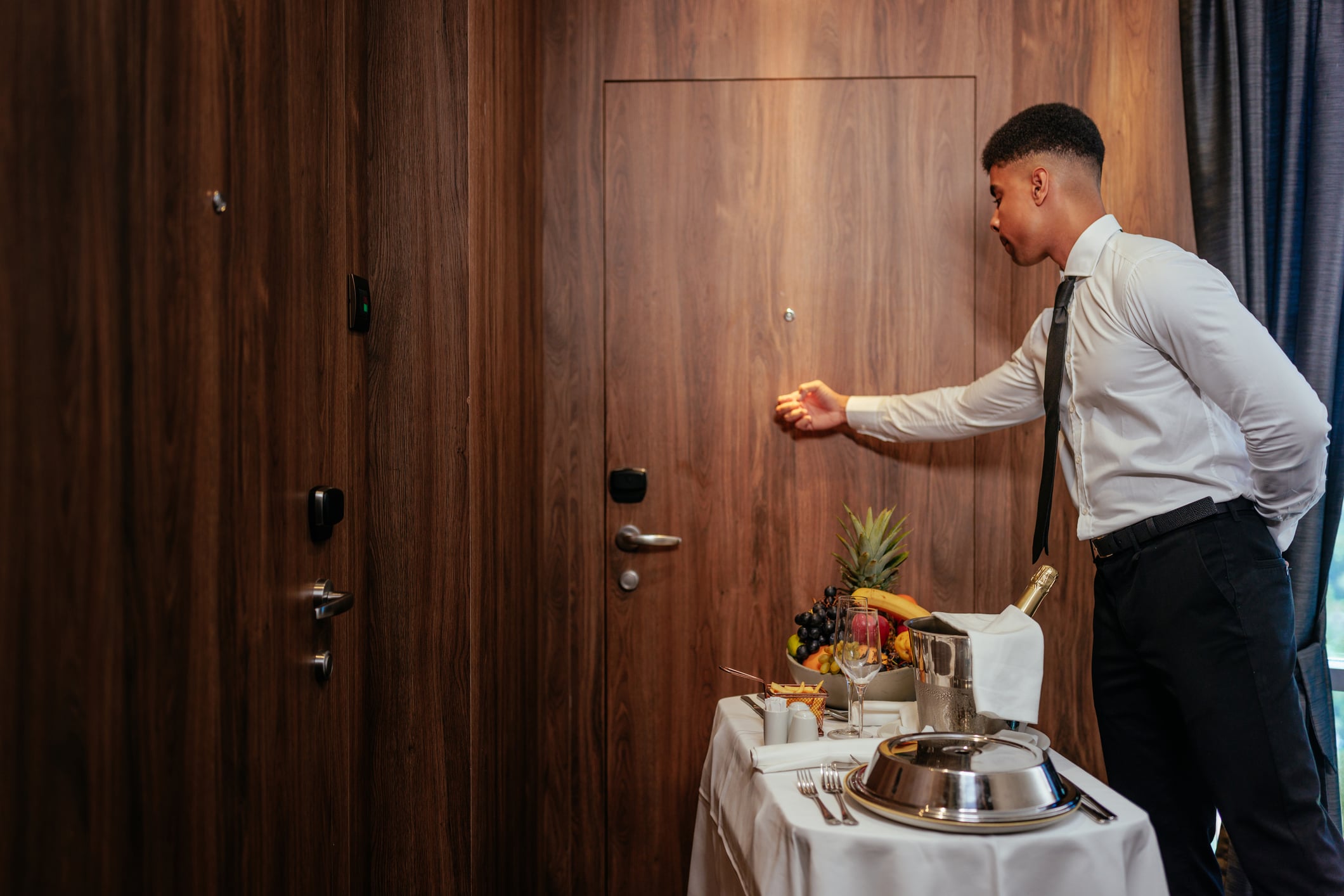 A hotel server in a white shirt and black tie knocks on a wooden door next to a room service cart with a fruit basket and a chilled bottle of champagne.