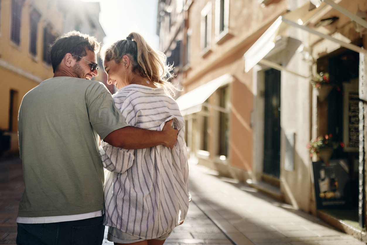 A couple walks arm in arm down a sunlit city street, looking at each other and laughing.