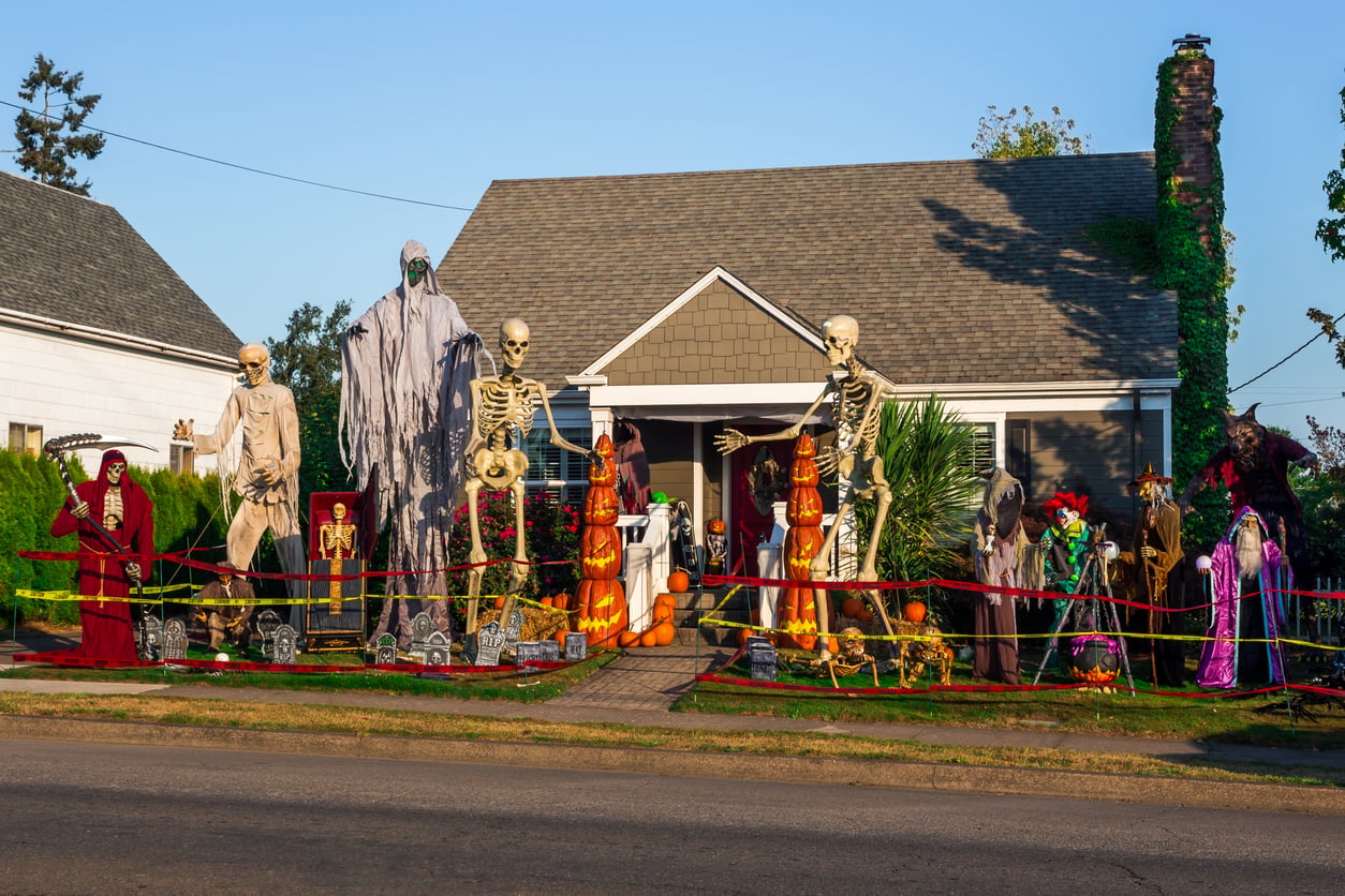 A residential front yard decorated for Halloween with giant skeletons, a ghostly figure, jack-o’-lanterns, and various monsters behind yellow caution tape.