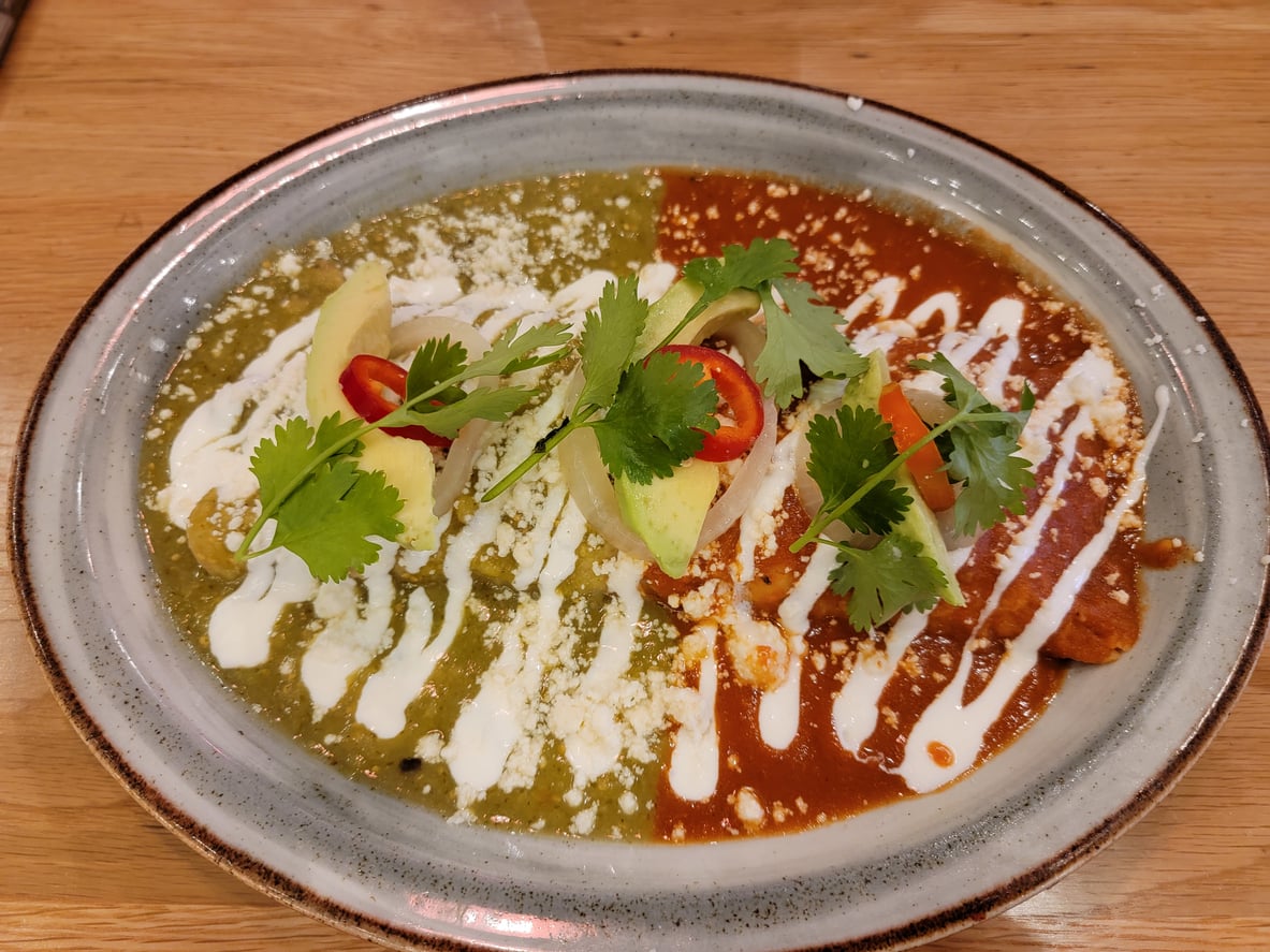 Enchiladas on a plate, half covered in green sauce and half in red sauce, topped with avocado, onion slices, chili peppers, cilantro, and drizzled with crema.