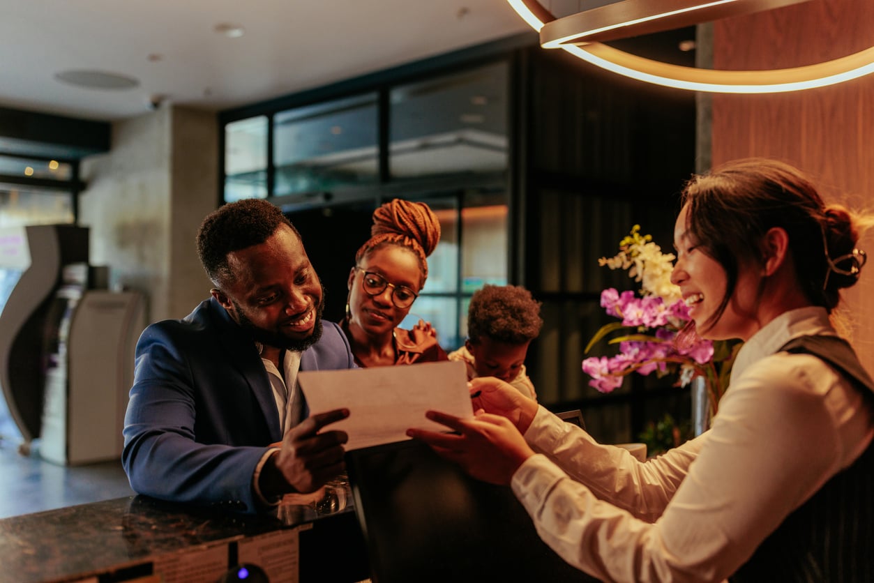 A smiling hotel receptionist hands a document to a family making trip planning decisions as they check in at the front desk.