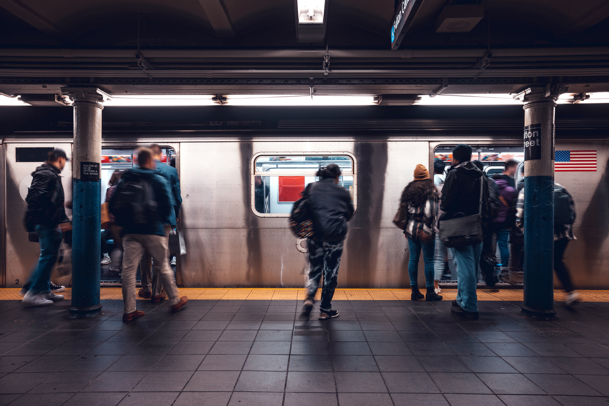 People wait on a subway platform as others board a silver train. The scene is indoors with fluorescent lights, blue columns, and a tiled floor. An American flag is visible on the train.