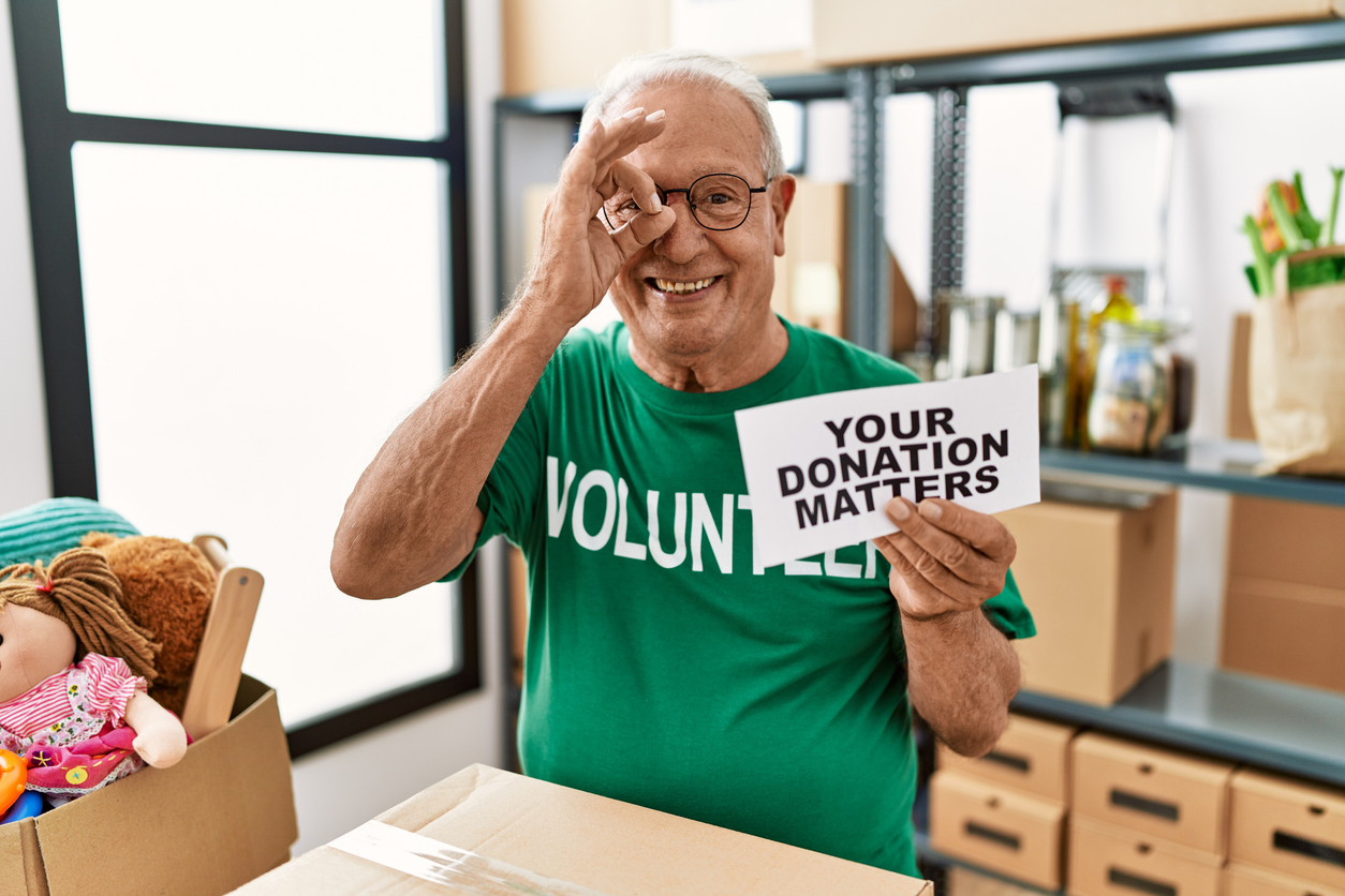 A smiling senior volunteer in a green t-shirt makes an okay gesture with his hand over his eye while holding a sign that says, “YOUR DONATION MATTERS,” spending time in a charitable cause.