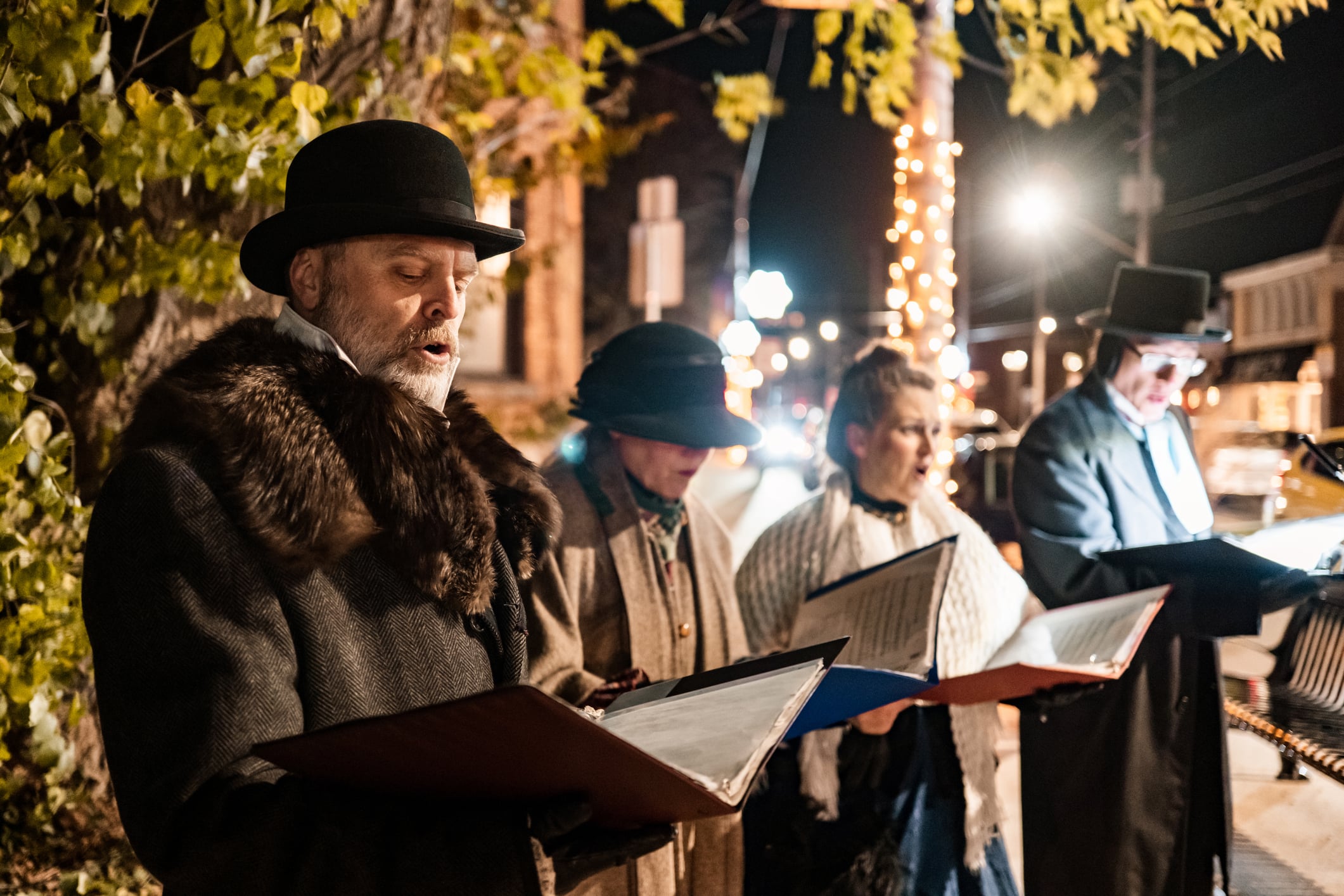 Four people in period-appropriate winter clothing, including top hats and coats with fur collars, singing from songbooks outdoors at night.
