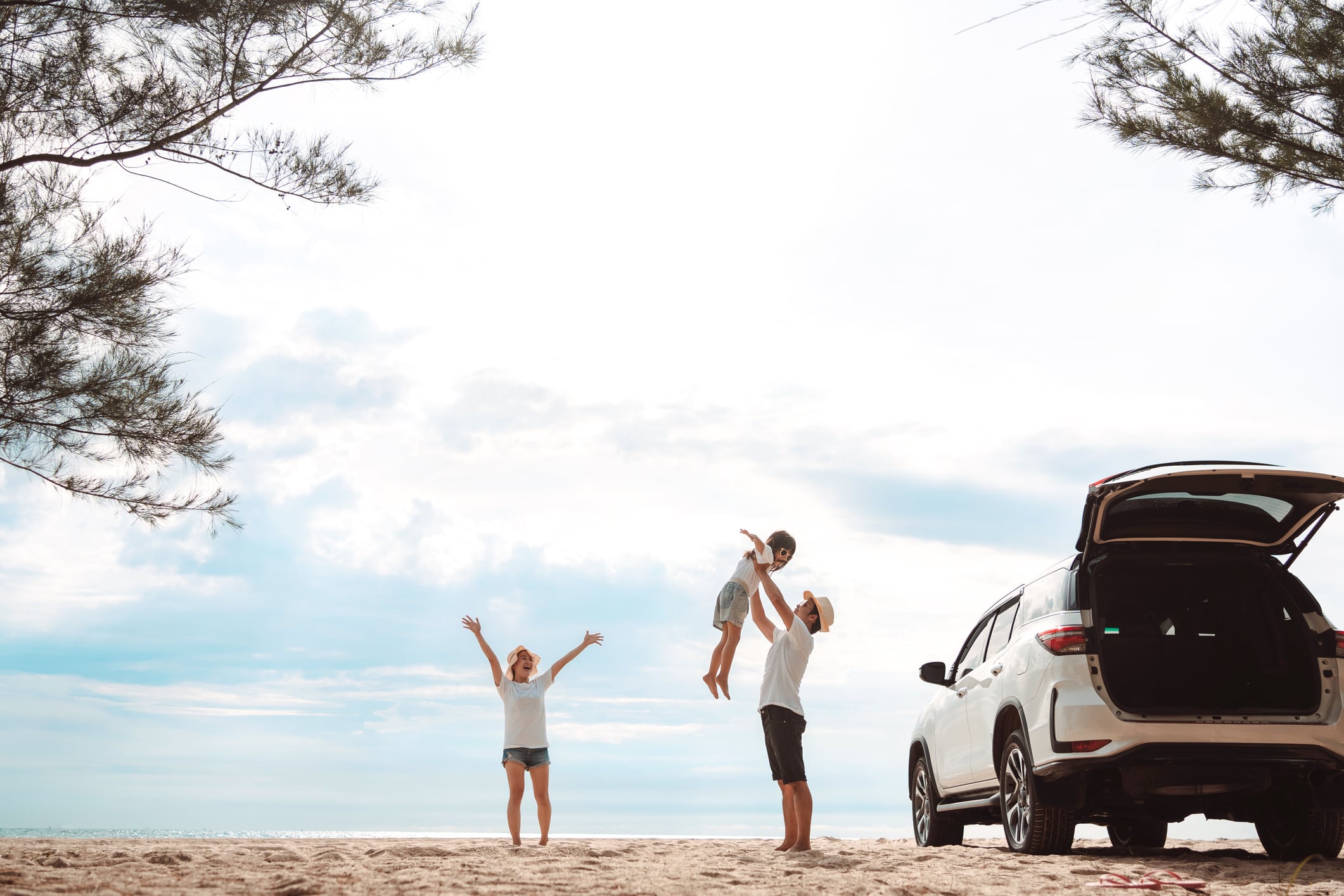 A father lifts his child into the air while a woman stands nearby with her arms raised on a beach next to an SUV with its trunk open.