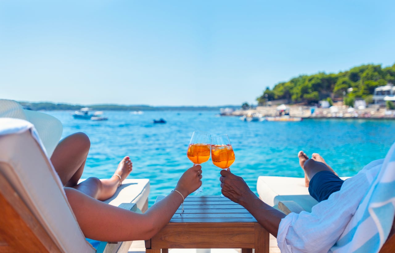 Two people lounging on deck chairs by the sea toast each other with orange cocktails.
