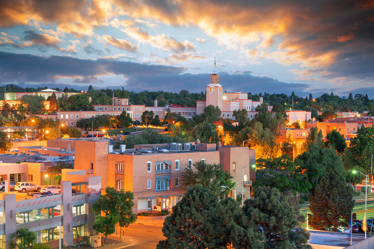 Downtown Santa Fe, New Mexico at sunset, with adobe-style buildings, green trees, and dramatic clouds lit by the setting sun, creating a vibrant and warm cityscape.