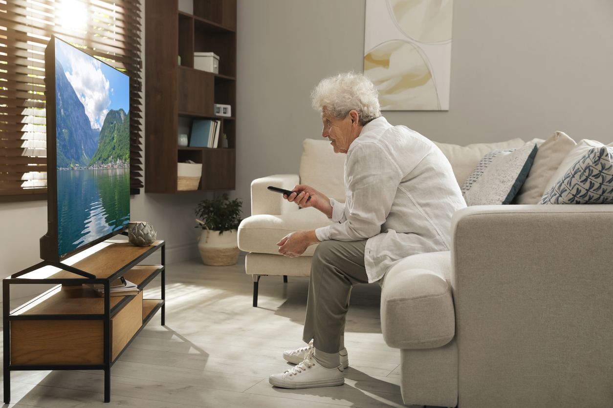 An elderly woman sitting on a couch points a remote control at a television displaying a landscape of mountains and a lake.