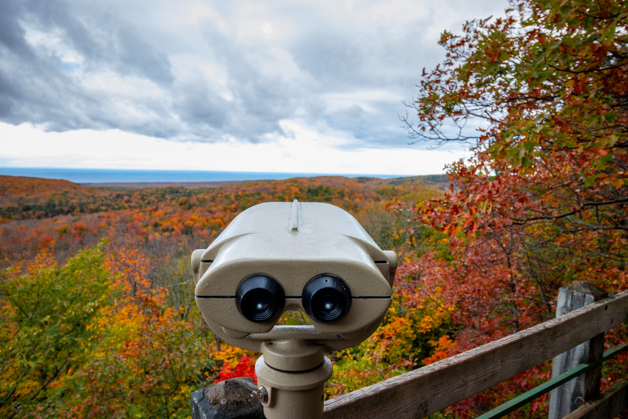 Stationary tourist binoculars overlooking one of the many places to hike in the fall, featuring a forest with autumn foliage under a cloudy sky.