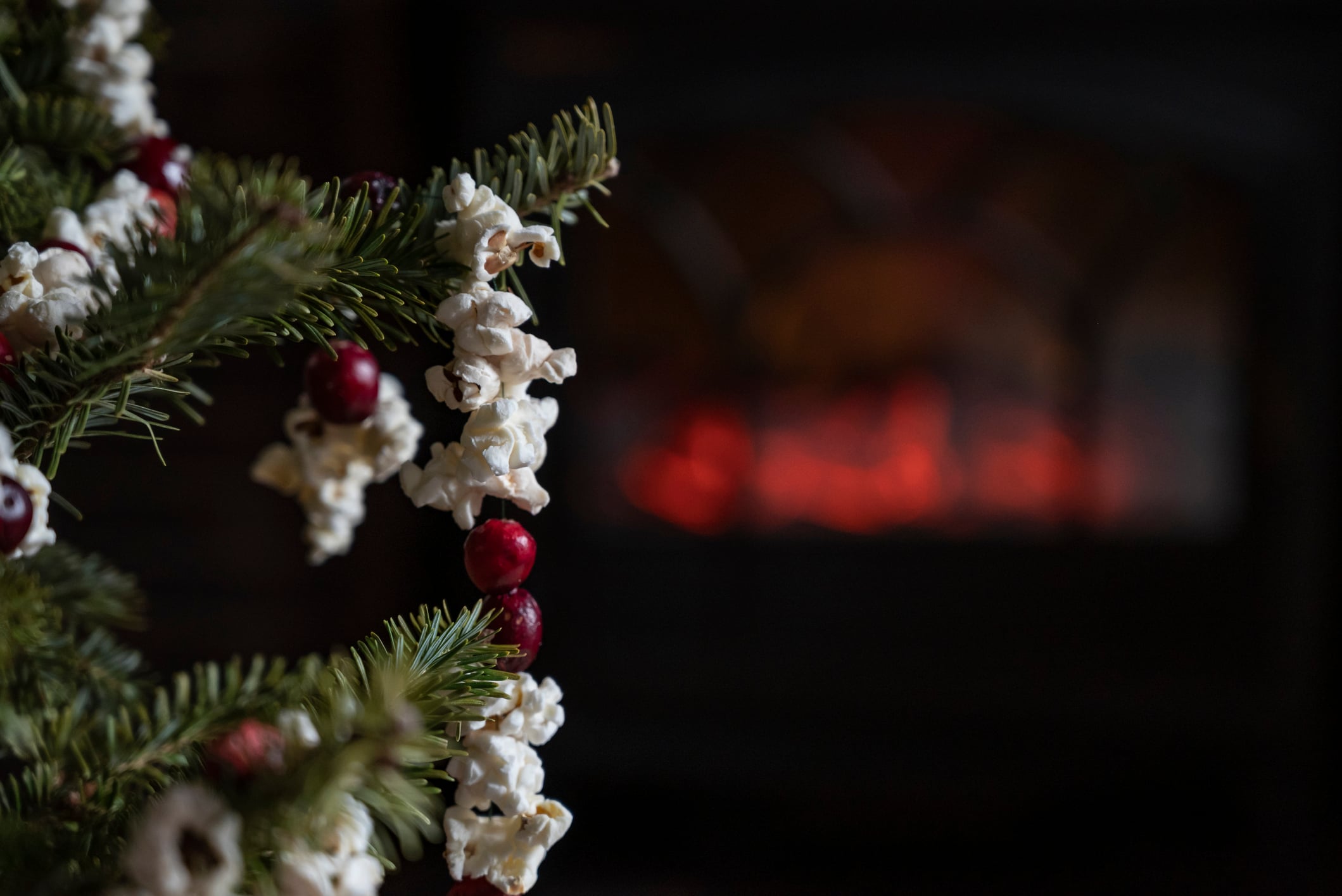 A string of popcorn and cranberries decorating a Christmas tree branch, with a glowing fireplace blurred in the background.