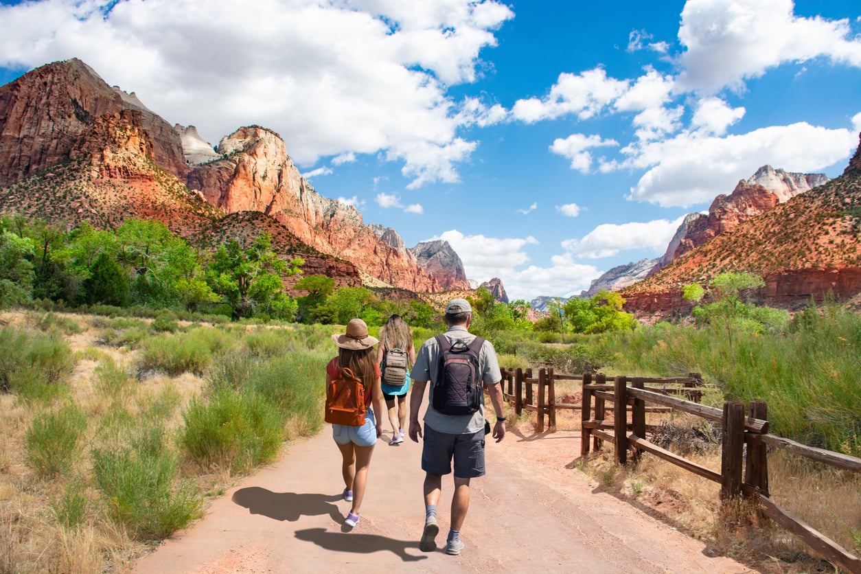 Three people with backpacks walk along a dirt path in a valley surrounded by rocky red mountains.