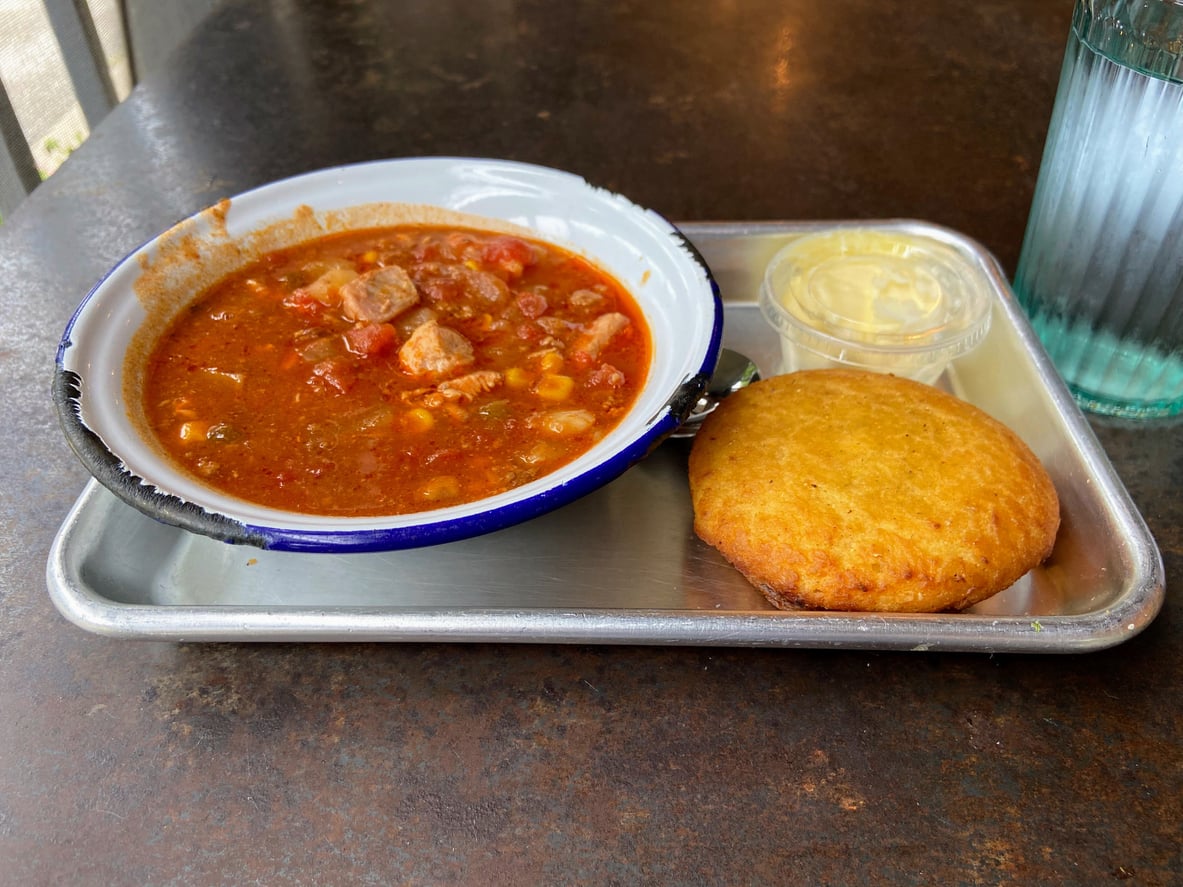 A bowl of chunky red stew, a round fried corn cake, and a small container of butter on a metal tray.