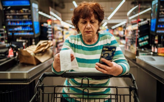 A woman in a striped sweater holds a long paper receipt and a smartphone while standing at a grocery store checkout.