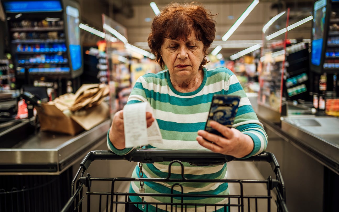 A woman in a striped sweater holds a long paper receipt and a smartphone while standing at a grocery store checkout.