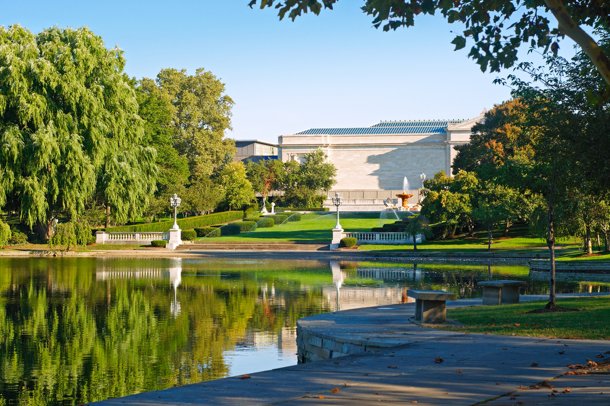 A stone walkway edges a pond reflecting a landscaped park with a fountain and a large, neoclassical museum building in the distance.