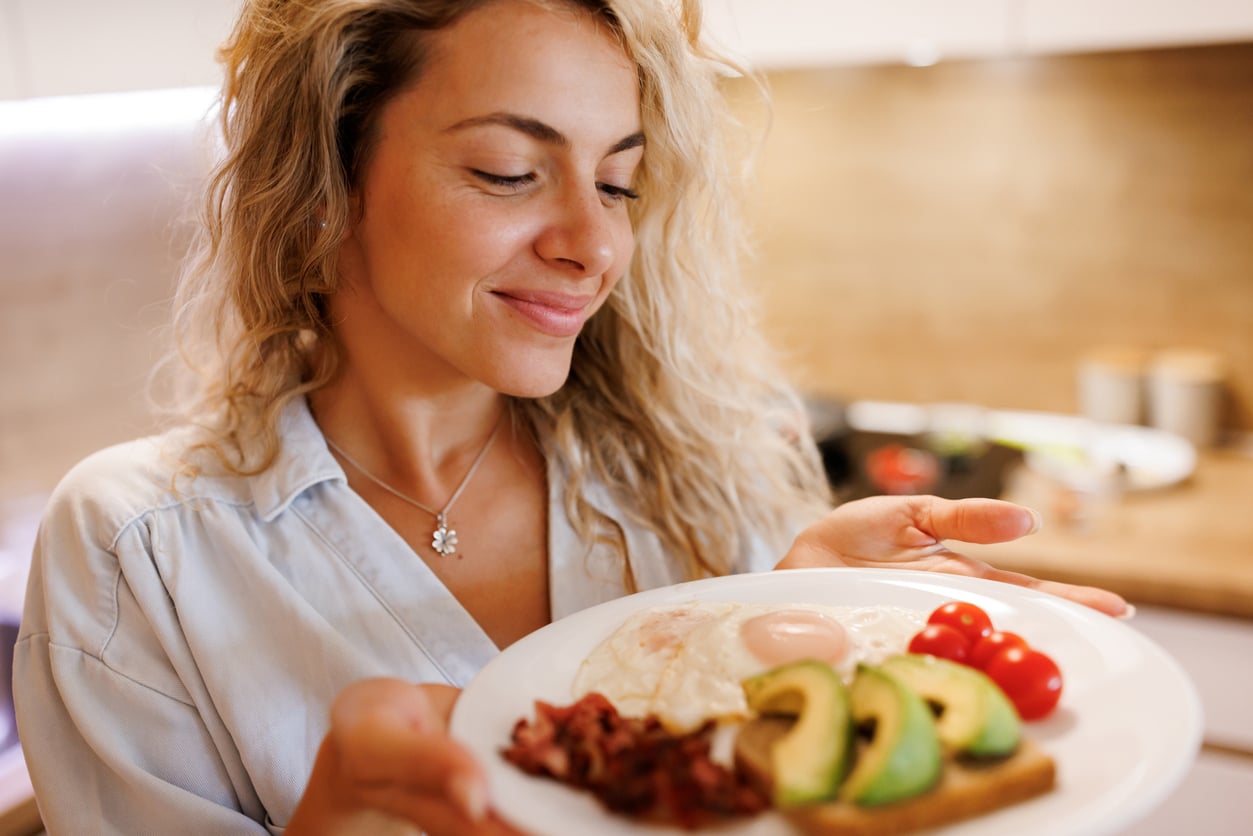 A smiling woman holding a plate of fried eggs, bacon, avocado slices, and cherry tomatoes.
