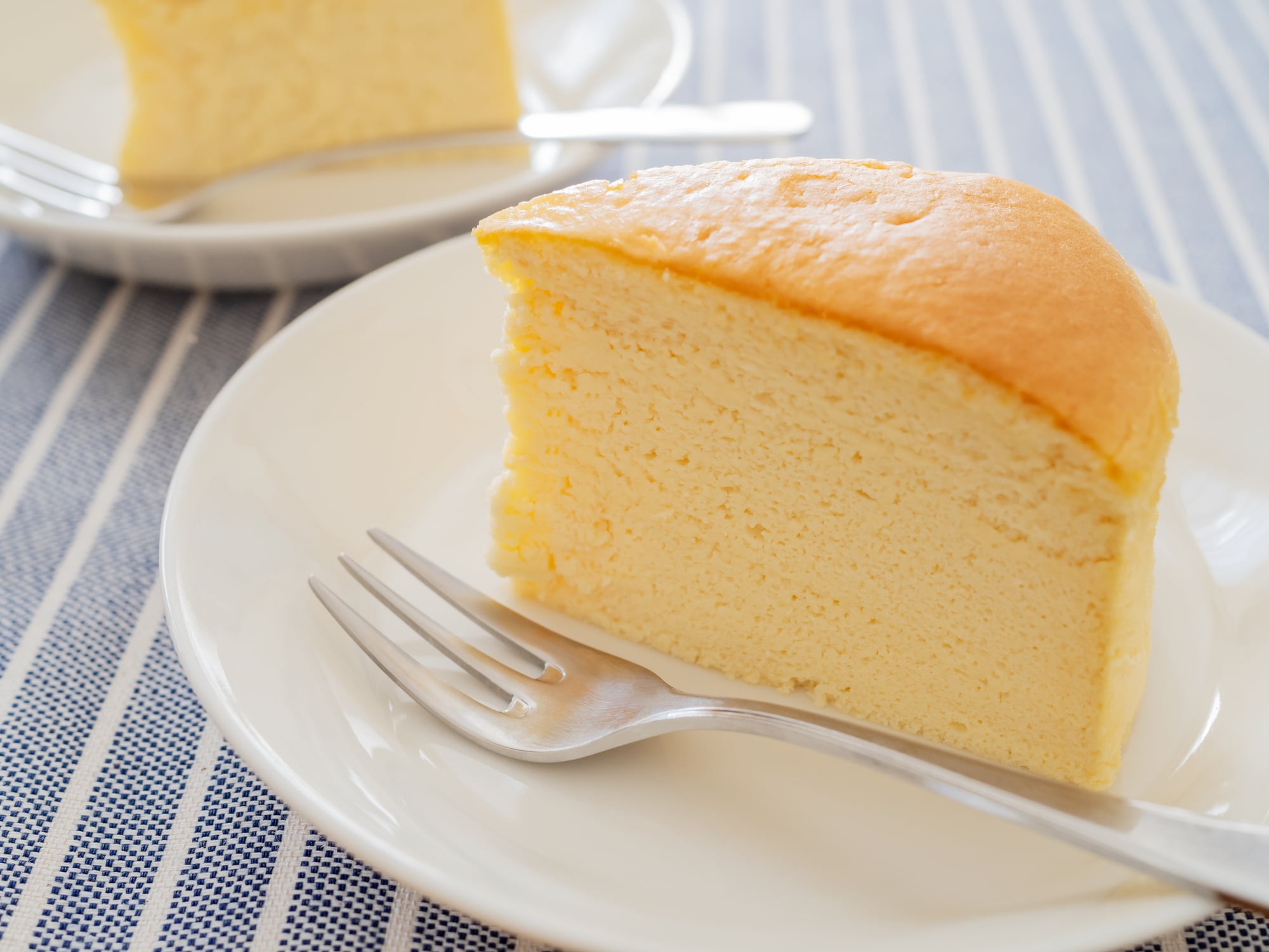 A slice of Japanese cheesecake on a white plate with a fork, with another slice visible in the background.