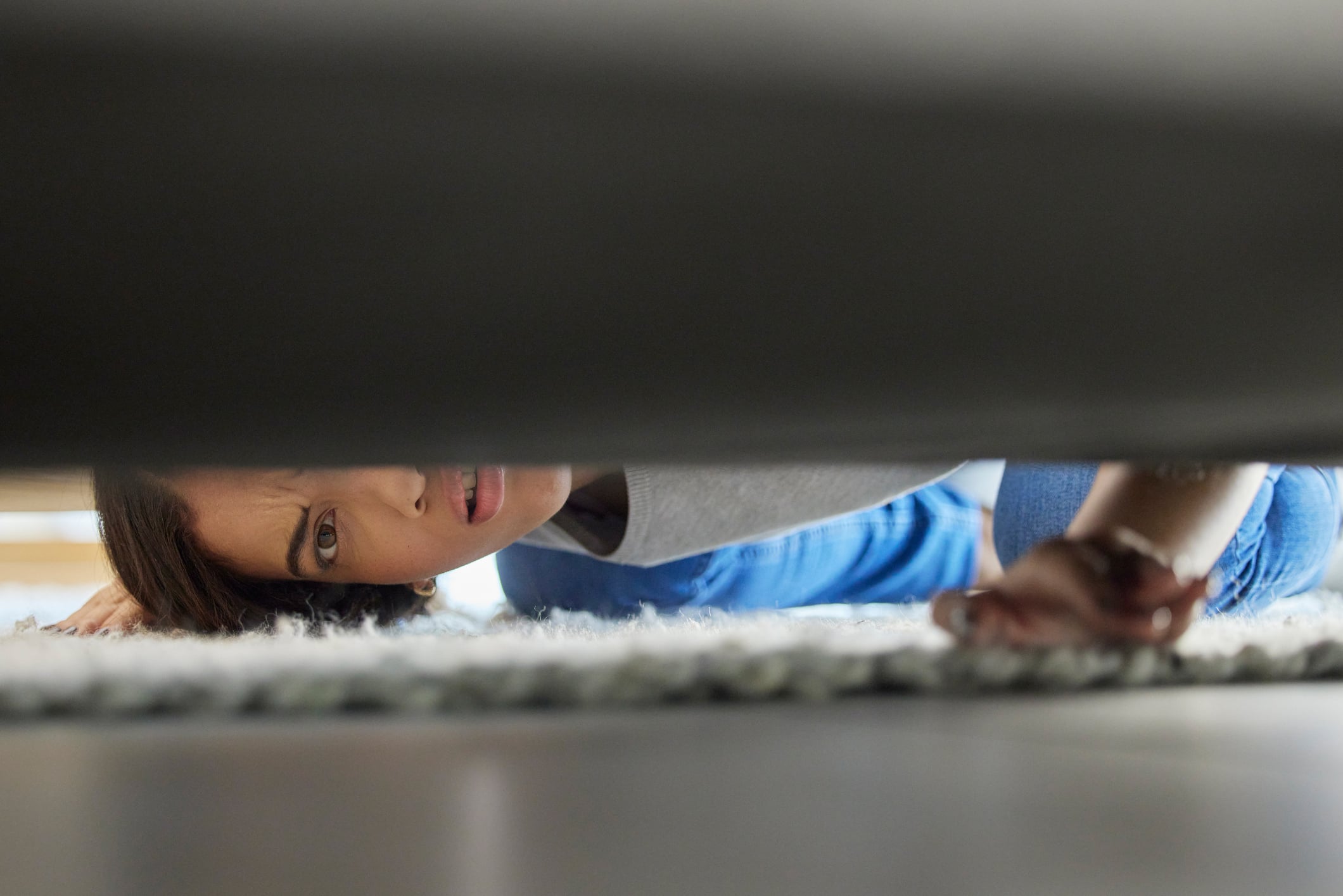 A woman lies on a rug peering underneath a low piece of furniture while working on home improvements.