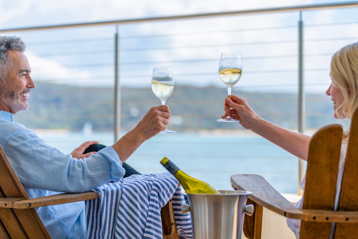 A man and woman sit in wooden chairs on a balcony overlooking the water, toasting each other with glasses of white wine.